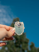 Hand holding a sticker with a book design against a blue sky