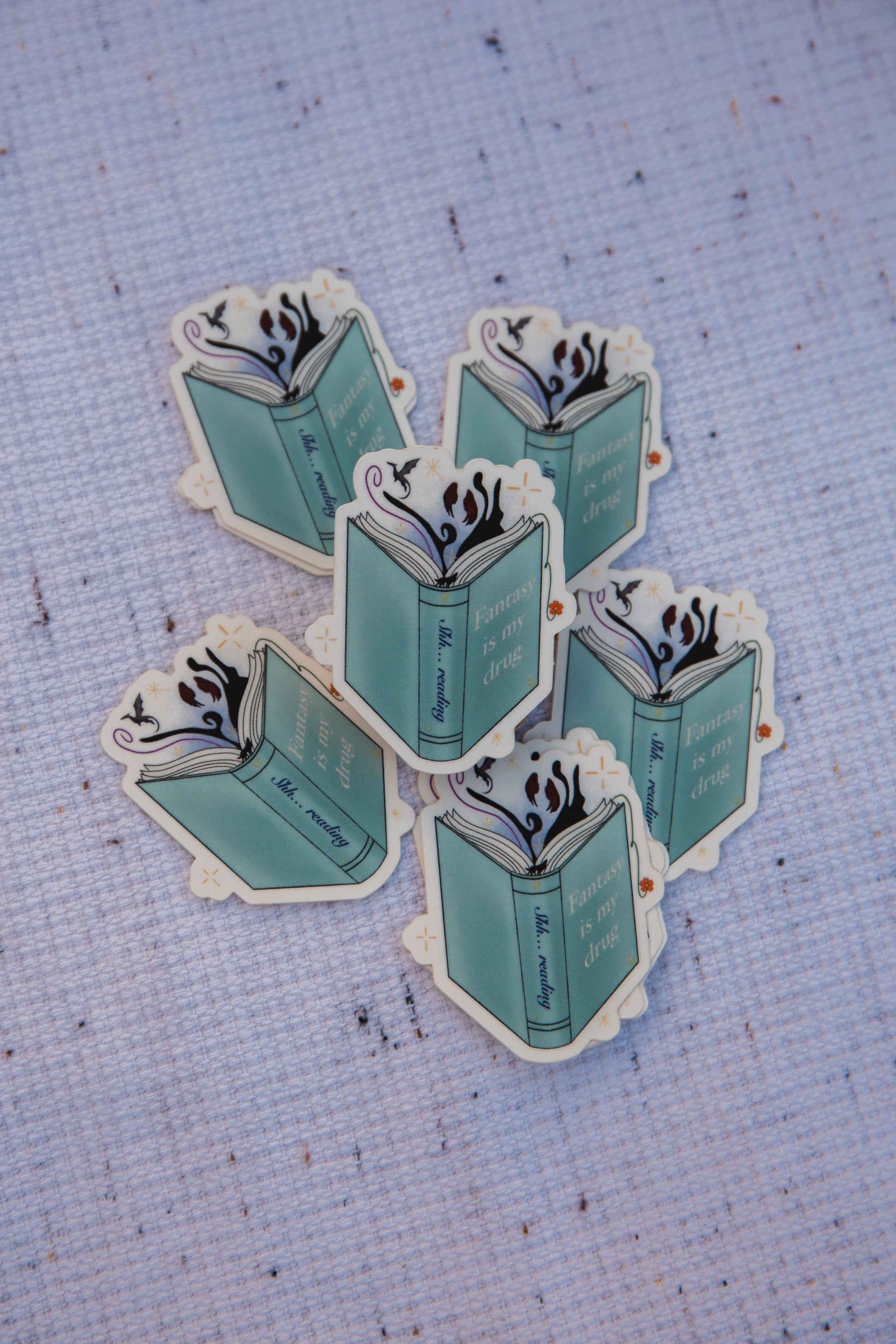 six books stickers layer out on top of a woven cloth. the books are blue and have the writing " fantasy is my drug". there are also fantasy theme items coming out of the book