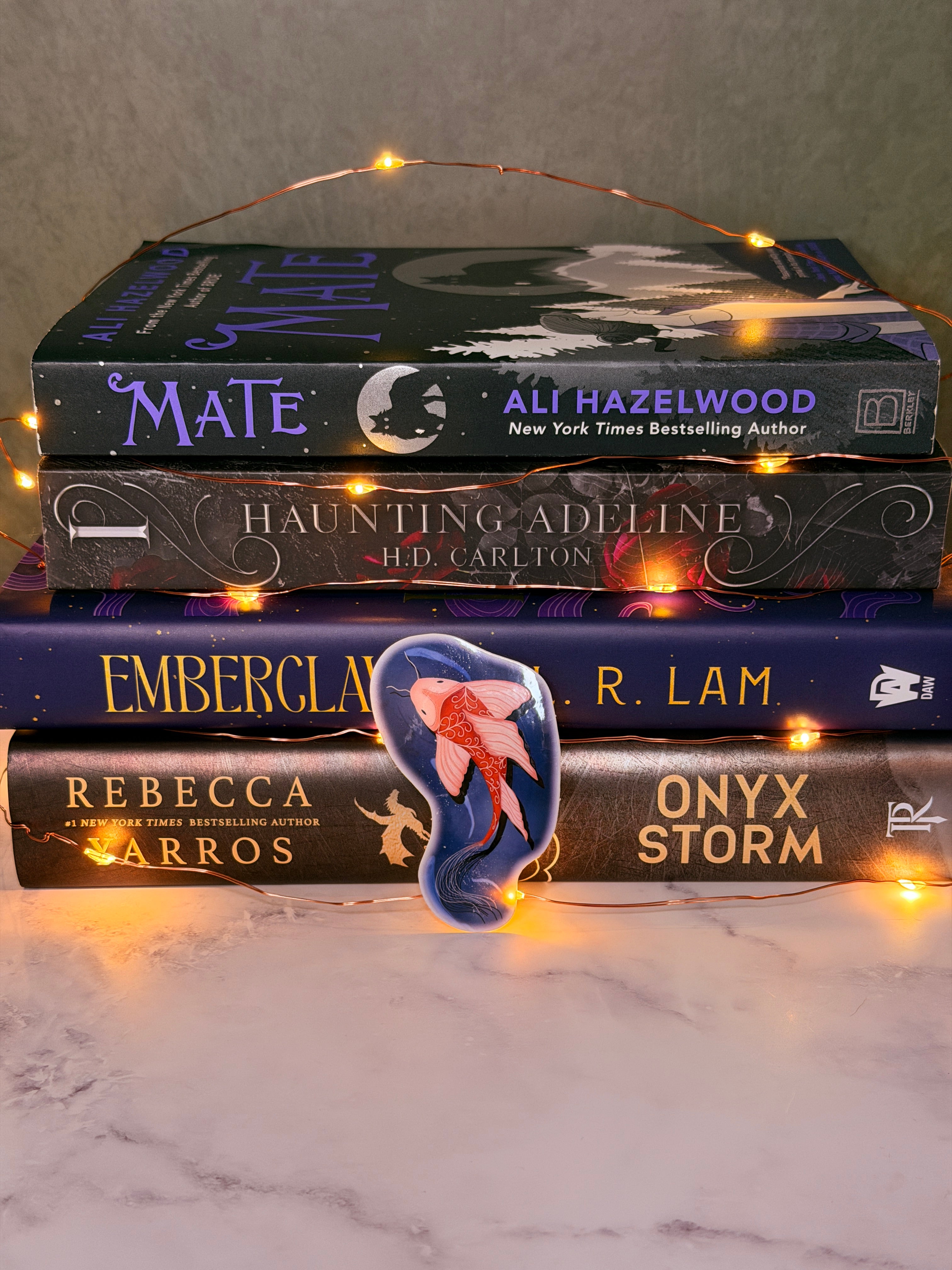 Four books titled 'Mate', 'The Haunting of Hill House', 'Onyx Storm', and 'Emberclan' on a marble surface with string lights.