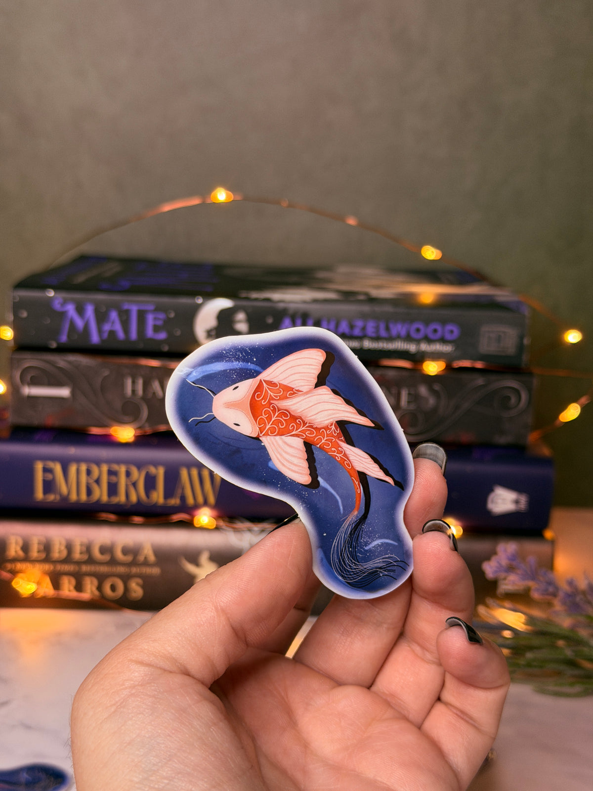 Hand holding a sticker of an orange fish swimming in dark blue water. With blurred books and lights in the background